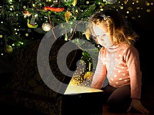 Child by Christmas tree opens magic chest. Portrait of little girl looking inside of gift box at home