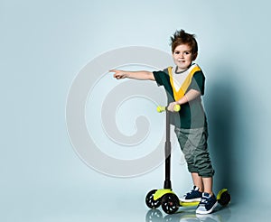 Child in colorful t-shirt, gray jeans, sneakers. He is pointing at something, standing on kick scooter on blue background
