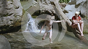 Child comes out of a small pond under a waterfall. CREATIVE. Mom watches as her son swims in a small mountain river near