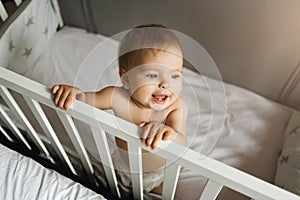 The child is in the crib. Portrait of a nine-month-old smiling baby girl standing in the playpen. Cheerful happy child