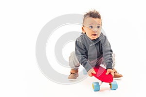 Child curiosity concept. Young multiracial baby boy playing with mini red skateboard in a studio over white background.