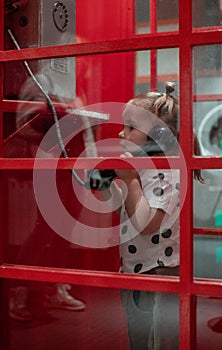 Child cute little girl in a red traditional phone booth. The little girl holds the handset of an old telephone in her