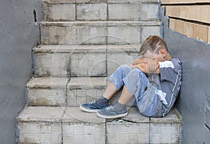 A child in depression sits on the stairs hugging his knees with his hands. In summer, a lonely boy is sad with his face