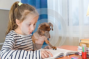 Child with dog reading book at home. Girl with pet sitting at window at read