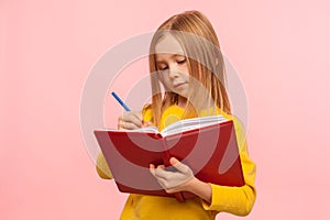 Child doing homework. Portrait of clever little girl writing poem in book with serious concentrated expression