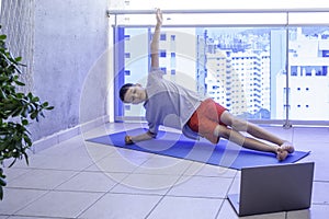 Child doing sport exercises side plank with laptop computer on balcony. Sport, healhty lifestyle, active leisure at home
