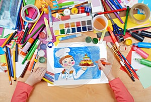 Child drawing holiday cake and cook, top view hands with pencil painting picture on paper, artwork workplace