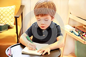 Child is drawing and painting with felt pen on paper of spiral notebook on small wooden table in living room at home. Childhood.