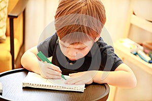 Child is drawing and painting with felt pen on paper of spiral notebook on small wooden table in living room at home. Childhood.