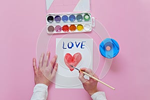 The child draws a card for Mother`s Day, uses a brush and watercolor paints.