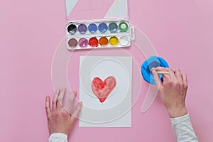 The child draws a card for Mother`s Day, uses a brush and watercolor paints.