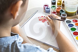 The child draws gouache on a white sheet and jars of paint are on the table. Hobbies and entertainment for children
