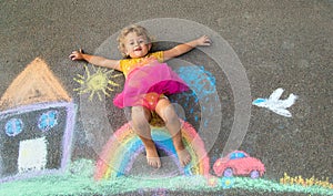 A child draws a house and a rainbow on the asphalt with chalk. Selective focus.