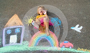 A child draws a house and a rainbow on the asphalt with chalk. Selective focus.