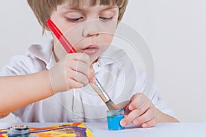 The child draws with paints. He puts a brush in blue paint