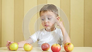 The child eats and plays with red and green apples, sitting at the table and showing the thumb of pleasure.