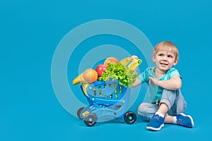 A child of European appearance, a blond boy, is sitting on the floor near a shopping trolley from a supermarket filled with
