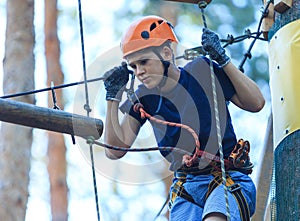 Child in forest adventure park. Kid in orange helmet and blue t shirt climbs on high rope trail.