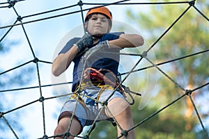 Child in forest adventure park. Kid in orange helmet and blue t shirt climbs on high rope trail.