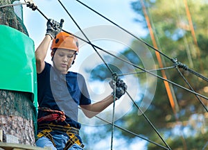 Child in forest adventure park. Kid in orange helmet and blue t shirt climbs on high rope trail.