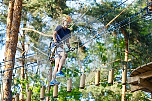 Child in forest adventure park. Kid in orange helmet and blue t shirt climbs on high rope trail.