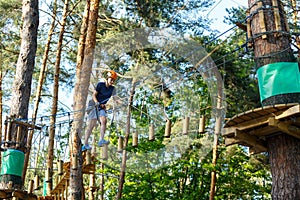 Child in forest adventure park. Kid in orange helmet and blue t shirt climbs on high rope trail.