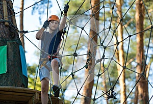 Child in forest adventure park. Kid in orange helmet and blue t shirt climbs on high rope trail.