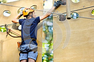 Child in forest adventure park. Kid in orange helmet and blue t shirt climbs on high rope trail. Agility skills