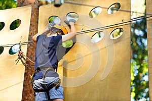 Child in forest adventure park. Kid in orange helmet and blue t shirt climbs on high rope trail. Agility skills