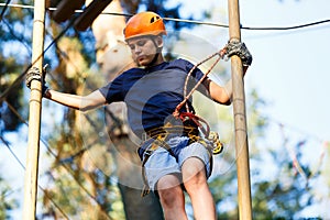 Child in forest adventure park. Kid in orange helmet and blue t shirt climbs on high rope trail. Agility skills