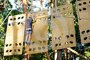 Child in forest adventure park. Kid in orange helmet and blue t shirt climbs on high rope trail. Agility skills