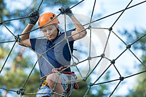 Child in forest adventure park. Kid in orange helmet and blue t shirt climbs on high rope trail. Agility skills