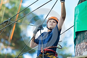 Child in forest adventure park. Kid in orange helmet and blue t shirt climbs on high rope trail. Agility skills