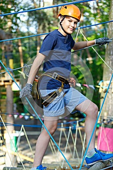 Child in forest adventure park. Kid in orange helmet and blue t shirt climbs on high rope trail. Agility skills