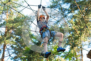 Child in forest adventure park. Kid in orange helmet and blue t shirt climbs on high rope trail. Agility skills