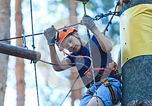 Child in forest adventure park. Kid in orange helmet and blue t shirt climbs on high rope trail. Agility skills and climbing