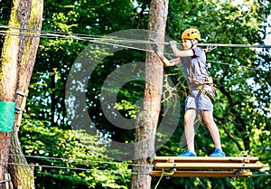 Child in forest adventure park. Kid in orange helmet and blue t shirt climbs on high rope trail. Agility skills and climbing