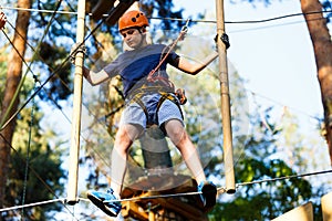 Child in forest adventure park. Kid in orange helmet and blue t shirt climbs on high rope trail. Agility skills and climbing