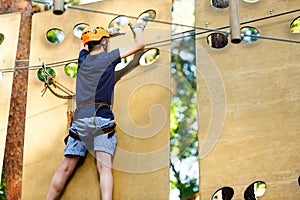 Child in forest adventure park. Kid in orange helmet and blue t shirt climbs on high rope trail. Agility skills and climbing