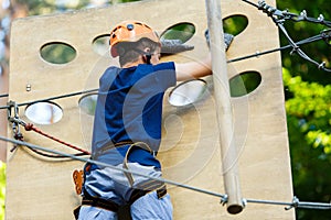 Child in forest adventure park. Kid in orange helmet and blue t shirt climbs on high rope trail. Agility skills