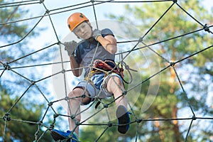 Child in forest adventure park. Kid in orange helmet and blue t shirt climbs on high rope trail. Agility skills