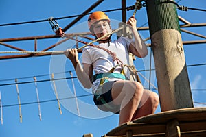 Child in forest adventure park. Kid in orange helmet and white t shirt climbs on high rope trail. Agility skills