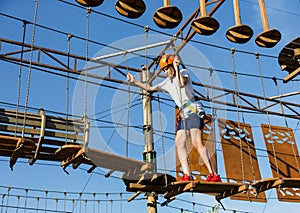 Child in forest adventure park. Kid in orange helmet and white t shirt climbs on high rope trail. Agility skills and climbing