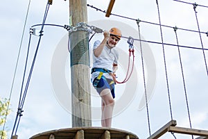 Child in forest adventure park. Kid in orange helmet and white t shirt climbs on high rope trail. Agility skills and climbing