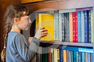 The child gets a textbook with wooden shelf of the old library. A little girl is taking a yellow book from a bookshelf. Children`