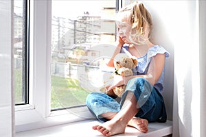 Child is a girl alone at home. Girls sitting on the windowsill and crying