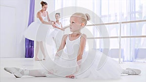 Child girl ballerina in white tutu is doing stretch exercises on ballet lesson.