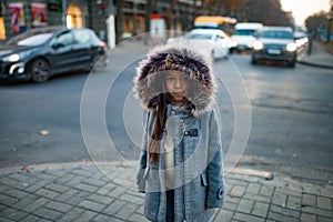 Child girl is standing at the sidewalk on background of city street in evening.