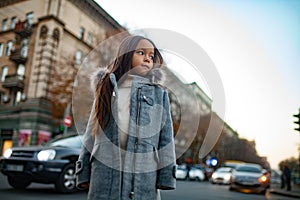 Child girl is standing at the sidewalk on background of city street in evening.