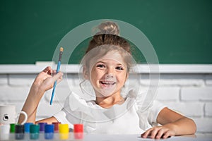 Child girl draws in classroom sitting at a table, having fun at school blackboard background. Painting school lesson